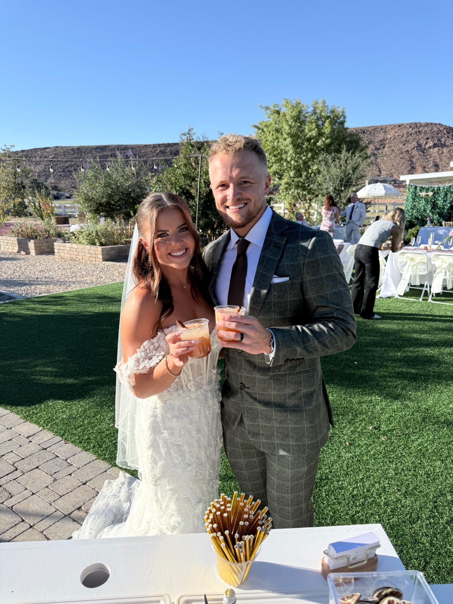 Two people in wedding attire smiling and holding garnished drinks beside the outdoor bar at Mckayla and Austin&rsquo;s Wedding