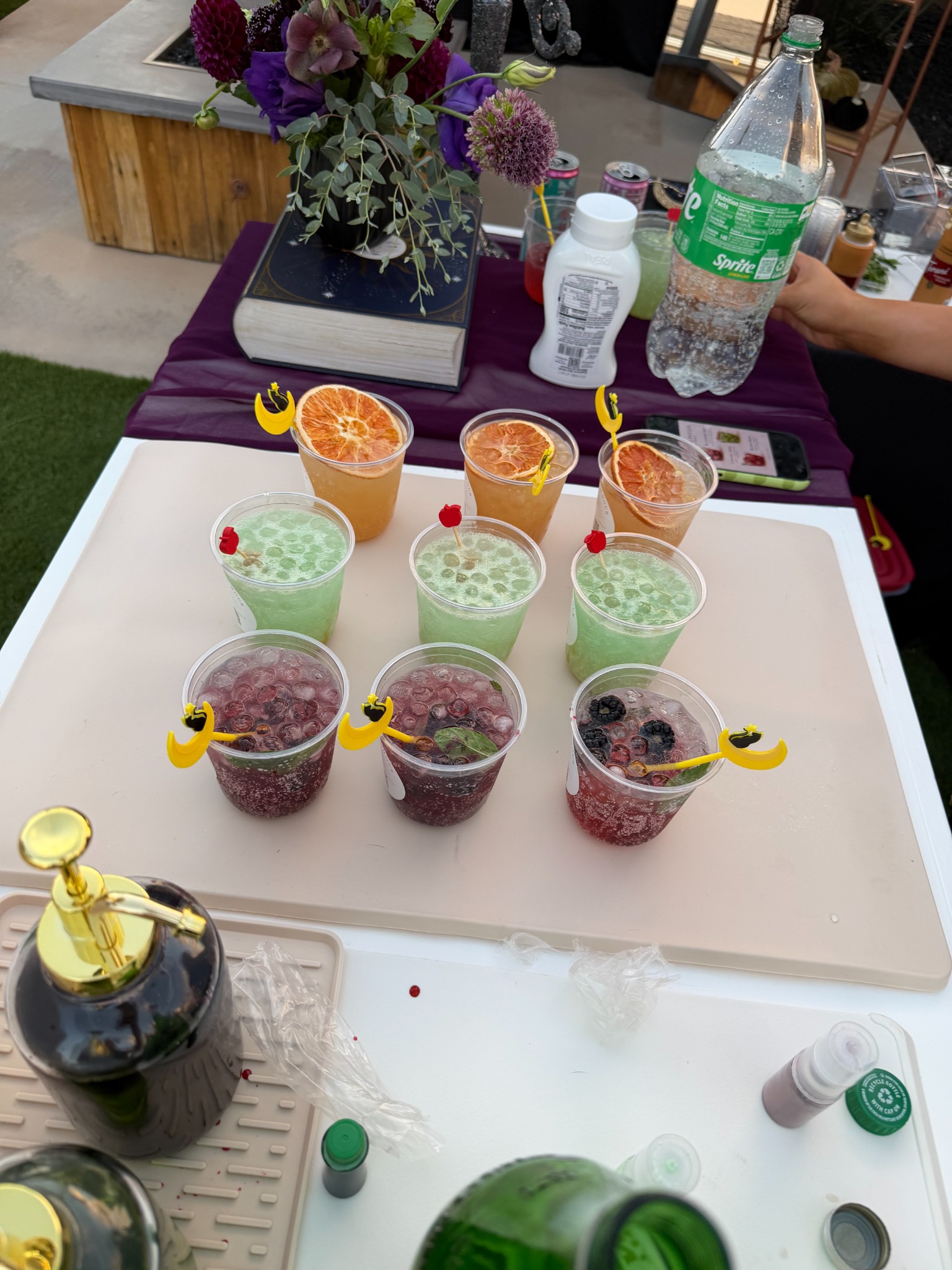 Nine iced drinks arranged in three colorful rows on the white bar with purple flowers and drink supplies behind them