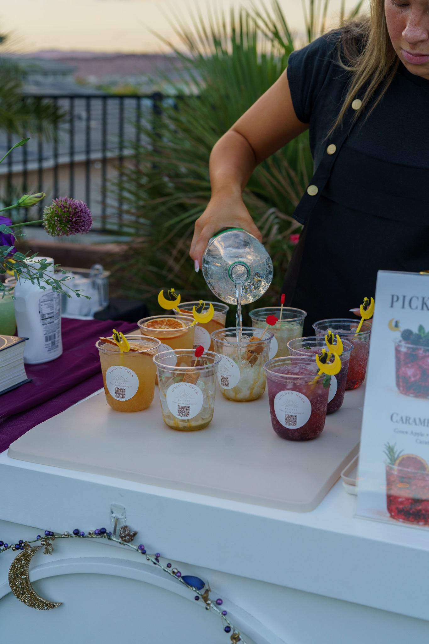 A server pours clear soda into a cup beside rows of colorful drinks on the white mobile bar