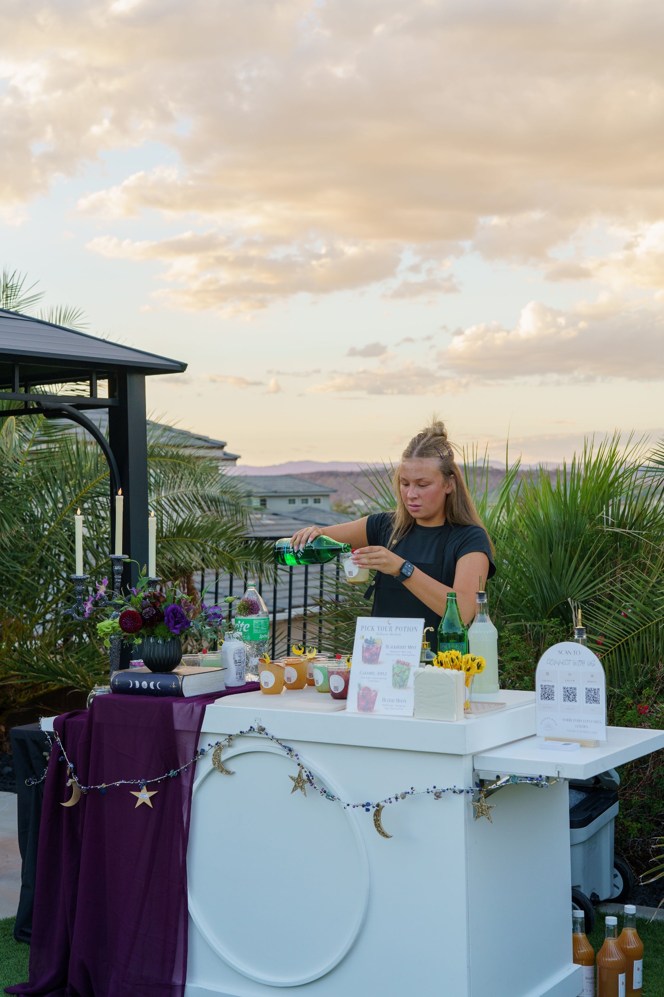 A server pours a drink behind the decorated Golden Hour Events cart beneath a cloudy sunset sky