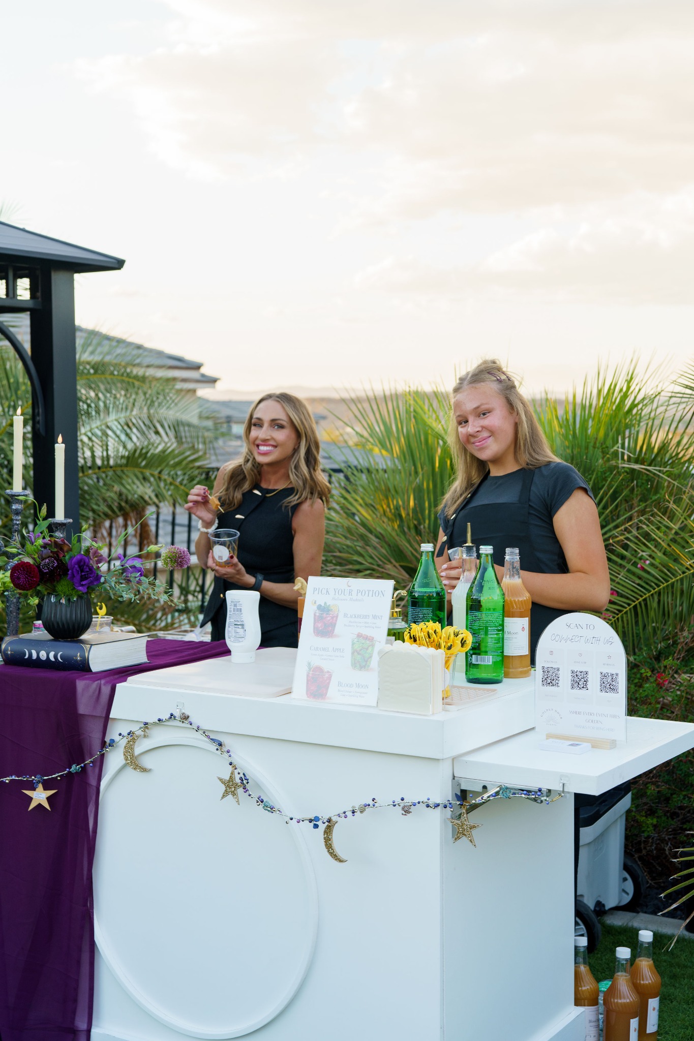 Two people dressed in black behind a white drink cart decorated with purple fabric, flowers, gold stars and crescent moons